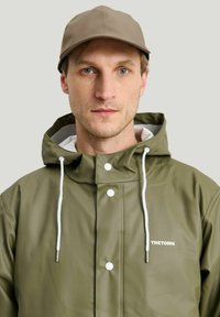 Grön vattentät jacka med vita dragsnören och knappar; har en hög krage och logga; bärs med en matchande beige keps.