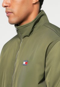 Ελαιόχρωμο μπουφάν με φερμουάρ από λείο ύφασμα, με ψηλό γιακά και ένα μικρό, πολύχρωμο λογότυπο της Tommy Jeans στο αριστερό μέρος του στήθους.