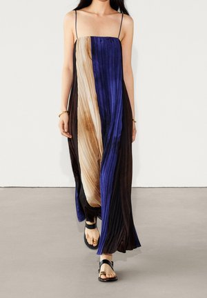 Vrouw draagt een mouwloze, geplooide maxi-jurk met verticale panelen in zwart, beige en blauw, gecombineerd met zwarte sandalen, lopend naar voren.