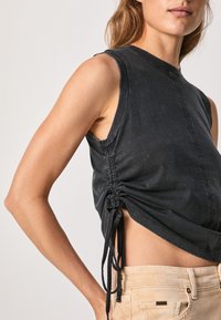 Svart ärmlös topp med rundad krage, texturerad tyg, sidofållning med band och en något lös passform, ihopparad med ljus beige byxor.