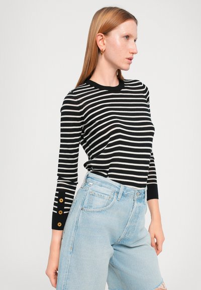 Maglietta a maniche lunghe a righe bianche e nere con scollo rotondo e bottoni decorativi sulle maniche, abbinata a pantaloni corti oversize di colore blu chiaro.
