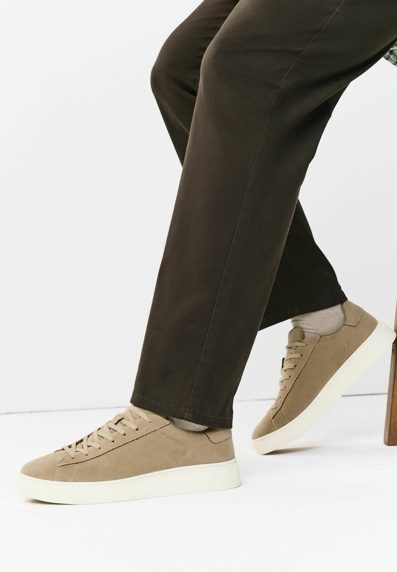 Jambes en pantalon sombre portant des baskets beiges en daim à lacets avec semelles blanches, assises devant un fond blanc uni.