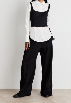 Woman wearing a white button-up shirt under a black cropped tank top with wide-leg black trousers and black shoes.