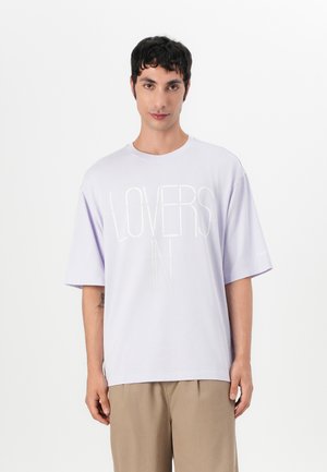 Lilla oversized t-shirt med korte ærmer, der har teksten "LOVERS IN" i en subtil, lysere nuance. Blødt bomuldsstof med en afslappet pasform.