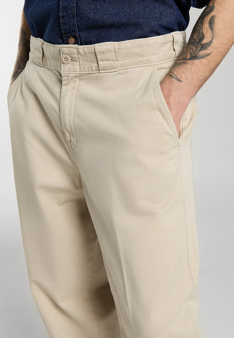 Uomo con pantaloni beige con le mani nelle tasche, che mostra braccia parzialmente tatuate e camicia blu scuro infilata.