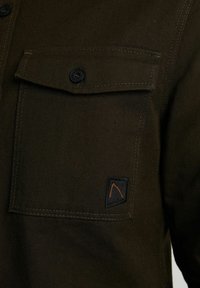 Ciemnooliwkowa koszula z guzikową kieszenią na piersi, z czarną metką z pomarańczowym emblematem. Teksturowana tkanina z widocznym przeszyciem.