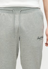 Κοντινό πλάνο γκρι φόρμας με ελαστική μέση με κορδόνι και μικρό μαύρο λογότυπο στο δεξί μηρό, φορεμένη με λευκό τοπ.