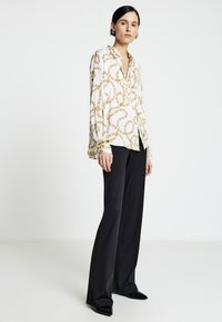 Blusa bianca a maniche lunghe con motivo a catena dorata, abbinata a pantaloni neri svasati e scarpe nere lucide.