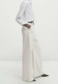 Chemise blanche courte boutonnée avec un ourlet festonné, associée à un pantalon crème taille haute à jambes larges et des talons noirs pointus.