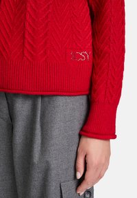 Maglione rosso lavorato a maglia con motivo a trecce, polsini a costine e una finitura texture. Decorato con la parola "SCSY" in accenti argentati.