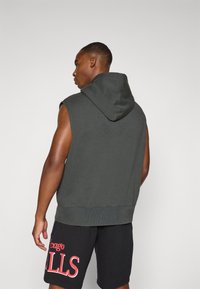 Sudadera sin mangas de color verde oscuro con capucha, hecha de una tela suave. Presenta un dobladillo acanalado y un diseño en la parte posterior. Combinada con pantalones cortos negros.