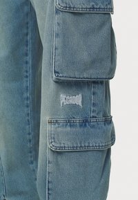 Svetlo modrá riflová bunda s viacerými náprsnými vreckami, štruktúrovanou tkaninou a vyšitou nášivkou "Pecador" v bielej farbe na hrudi.