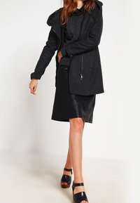 Zwarte wollen blendjas met een capuchon en rits, gedragen boven een donkere gebreide trui, gecombineerd met een zwarte leren rok en chunky sandalen.