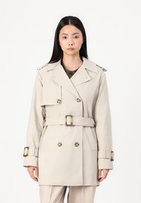 Beige trenchcoat med doble knapper, belte i livet og store knapper. Har skulderepauletter og brede revers. Glatt tekstur.