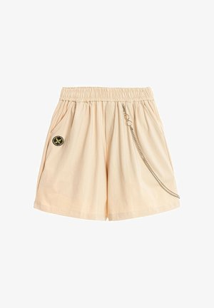 Pantalones cortos beige con cintura elástica, bolsillos laterales, un parche circular negro y amarillo en el lado izquierdo, y una cadena plateada colgando en el lado derecho.