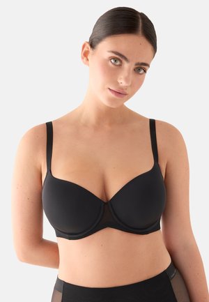 Mujer de cabello oscuro con un sujetador negro con aros y braguitas de talle alto a juego, posando contra un fondo gris claro.