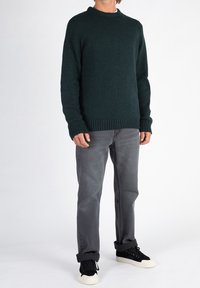 Man in een donkergroen gebreid vest, grijze opgerolde jeans en zwarte sneakers met witte zolen, staand op een witte achtergrond.