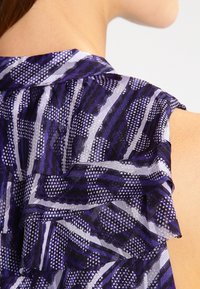 Fialová a bílá látka s vzorem a volánkovými okraji, přeložená přes rameno, vystavující složité geometrické vzory a texturu.