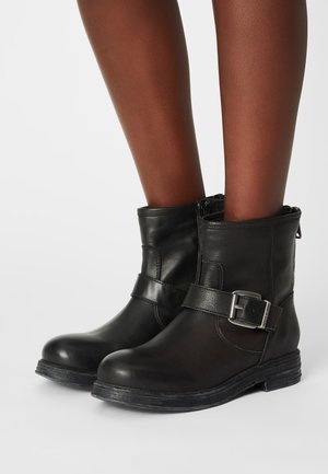 Botas de tornozelo em pele preta com biqueira arredondada, fecho lateral com zíper e uma faixa decorativa com fivela em metal. Sola em borracha, acabamento texturizado.