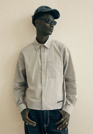 Junger Mann mit Sonnenbrille, einem grau karierten Hemd mit Knöpfen, einer dunklen Jeanskappe und dunklen Jeans, stehend vor einer schlichten hellen Wand.