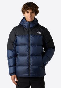 Puffjacka med marinblå och svarta färgblock, quiltat mönster, dragkedja fram och justerbar huva. Tillverkad av vattentåligt material.