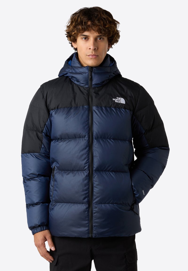 Puffjacka med marinblå och svarta färgblock, quiltat mönster, dragkedja fram och justerbar huva. Tillverkad av vattentåligt material.