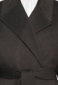 Abrigo de lana oscura con solapa ancha, diseño envolvente y un cinturón a juego en la cintura. Textura suave, detalles mínimos y silueta elegante.