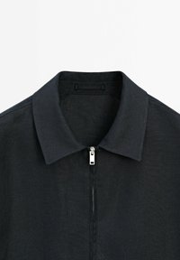 Veste noire à col avec fermeture éclair argentée centrée sous le col sur fond blanc uni.