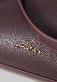 Sac à main bordeaux en cuir synthétique texturé, arborant un logo doré "ANNA FIELD" en relief à l'avant et une anse assortie.