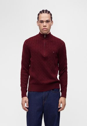Jeune homme aux cheveux tressés portant un pull bordeaux en maille torsadée avec fermeture éclair à moitié et un jean bleu foncé, debout devant un fond clair uni.