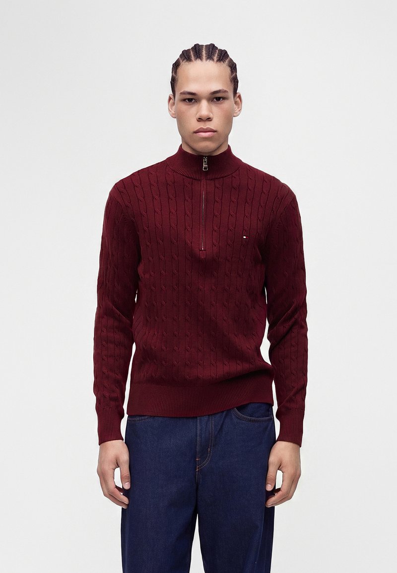 Jeune homme aux cheveux tressés portant un pull bordeaux en maille torsadée avec fermeture éclair à moitié et un jean bleu foncé, debout devant un fond clair uni.