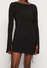 Robe noire à manches longues avec un décolleté bateau, dotée de liens sur les côtés à l'ourlet et d'une texture de tissu lisse et légèrement extensible.