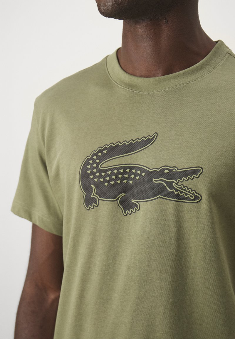 Olijfgroen katoenen T-shirt met een groot zwart krokodillenlogo met geometrische patronen. Klassieke ronde hals en ontwerp met korte mouwen.