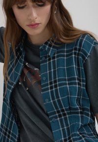 Camicia a quadri blu con un tessuto texturizzato, dotata di una chiusura con bottoni e dettagli neri, indossata sopra una t-shirt grigia con grafica.