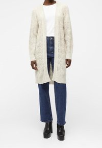 Cardigan beige clair en maille ajourée avec motif en torsades, manches longues et bordures côtelées, accompagné d'un jean bleu taille haute et de bottes à semelles compensées.