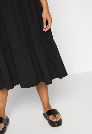 Schwarzes Baumwollkleid mit einem gestuften Design und einem Rüschensaum. Getragen mit klobigen schwarzen Sandalen, die einen dicken Riemen und eine Plattformsohle haben.