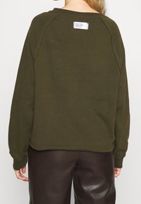 Sudadera de color verde oscuro con un ajuste relajado, mangas raglán y una etiqueta visible en la parte posterior. La tela parece suave y con textura.