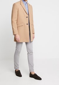 Tan wollen jas met twee knopen en gekartelde revers, gecombineerd met een blauw-wit geruit overhemd, grijze broek en donkerbruine instapschoenen.