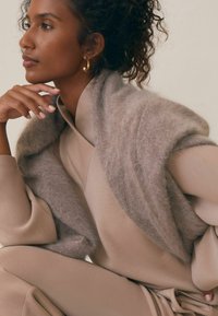 Beżowy, teksturowany, oversize szal nałożony na dopasowany beżowy strój, z szerokimi rękawami i talią podkreśloną paskiem. Widziane złote kolczyki w kształcie kół.