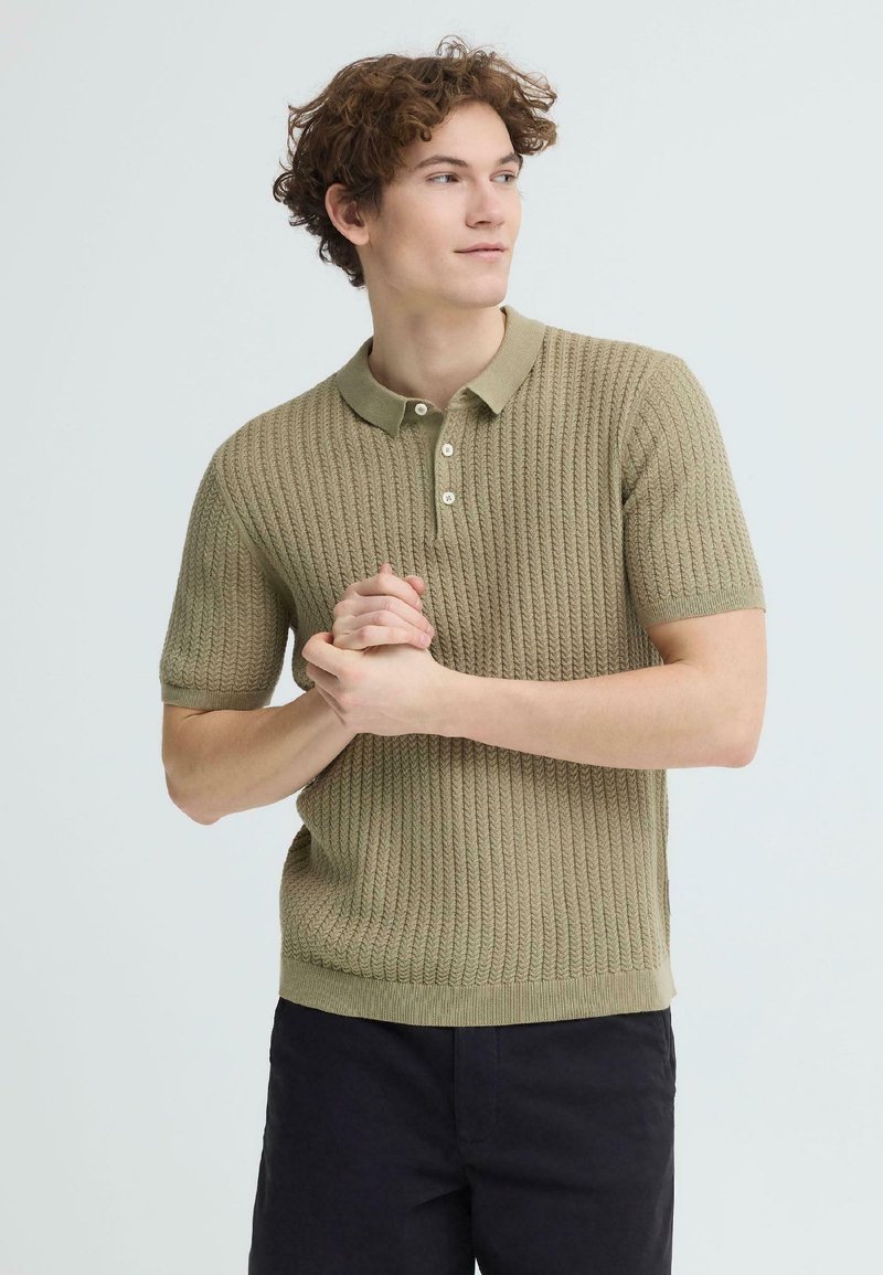 Νεαρός άνδρας με σγουρά μαλλιά, φορώντας ένα υφασμάτινο κοντομάνικο πόλο σε ελιά πράσινο και μαύρο παντελόνι, με τα χέρια ενωμένα, κοιτάζει προς τα δεξιά.