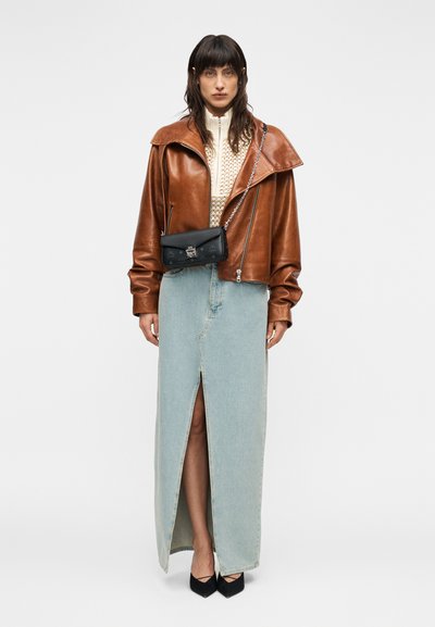 Γυναίκα με καφέ δερμάτινο μπουφάν, μπεζ πλεκτό πουλόβερ, φούστα από ανοιχτό μπλε τζιν με εμπρόσθια σκίσιμο, μαύρες μυτερές γόβες και μικρή μαύρη τσάντα ώμου.