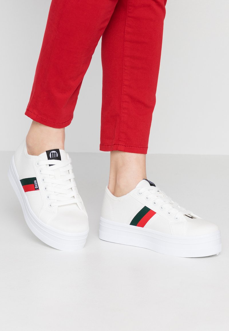 Baskets en toile blanches avec une semelle compensée, ornées d'une bande rouge et verte et d'un onglet logo noir, associées à un pantalon rouge cropped.