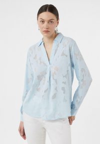 Hellblaues Langarmhemd mit kragenförmigem Ausschnitt, aus einem transparenten Stoff mit einem abstrakten Texturmuster. Weißer Hintergrund.