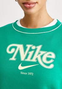 Grön sweatshirt i bomull, med en stor krämfärgad "Nike"-logga och en vit lagerkrage. Slät textur, avslappnad design.