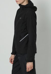 Persona che indossa una giacca nera con cappuccio, con una zip frontale e una striscia riflettente sul lato, in piedi di lato su uno sfondo semplice.