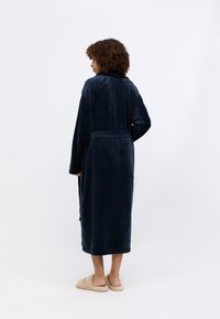 Tummansininen pörröinen aamutakki, jossa on pitkä ja virtaviivainen muotoilu. Varustettu huivikauluksella, sivutaskuilla ja sidottavalla vyöllä turvalliseen istuvuuteen.