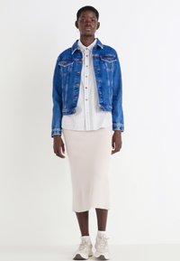 Veste en denim bleue, superposée à une chemise blanche à rayures avec un bouton. En dessous, une jupe crème ajustée, assortie à des baskets beiges.
