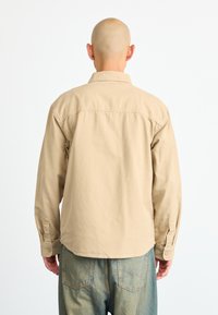 Camisa de manga larga beige claro con cuello, puños abotonados y detalles de costura sutiles. La tela tiene una textura suave.