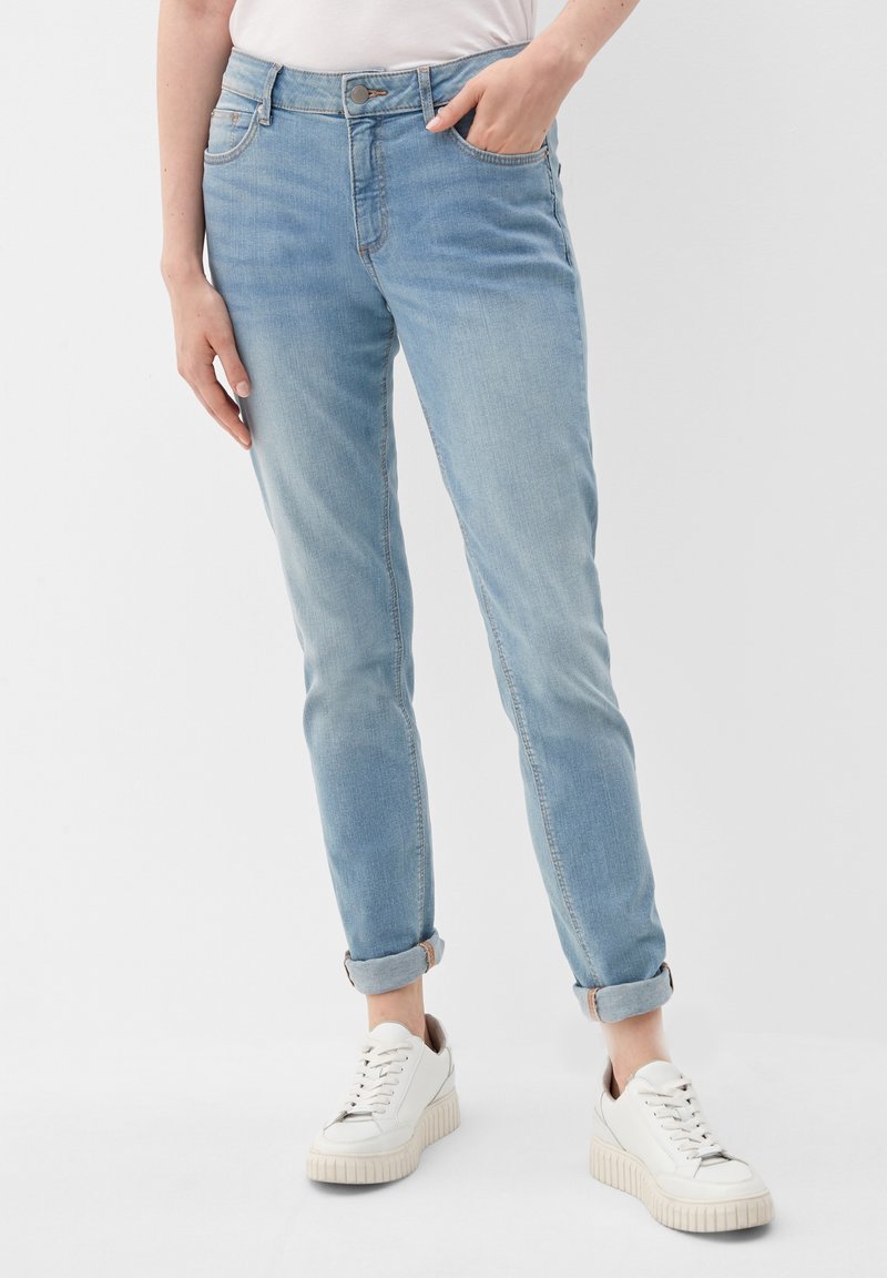 Personne portant un jean bleu clair retroussé et des baskets à plateforme blanches, avec une main dans la poche et l'autre bras détendu le long du corps.