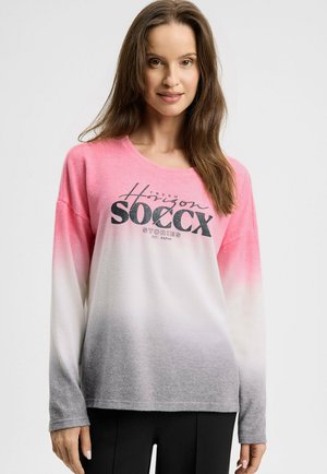Frau trägt einen langärmeligen Ombre-Sweatshirt in Pink, Weiß und Grau mit dem Aufdruck "Soccx" auf der Vorderseite, steht vor einem einfarbigen Hintergrund.
